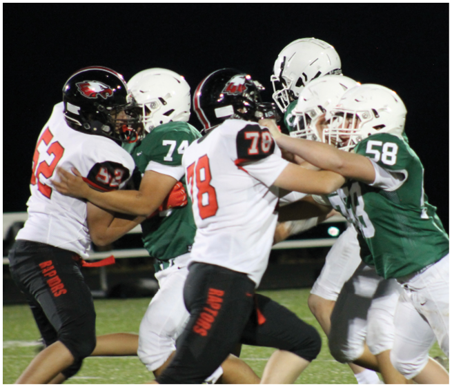 People wearing football uniforms in black and green are engaged in a blocking action on a football field at night. Description generated by AI
