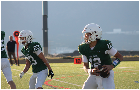 Two people wearing green football jerseys and white helmets are on a field, with one holding a football and the other preparing to move. Description generated by AI