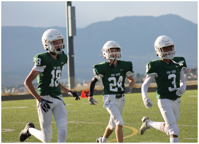 Three people wearing green football jerseys and white helmets are running on a football field with mountains in the background. Description generated by AI