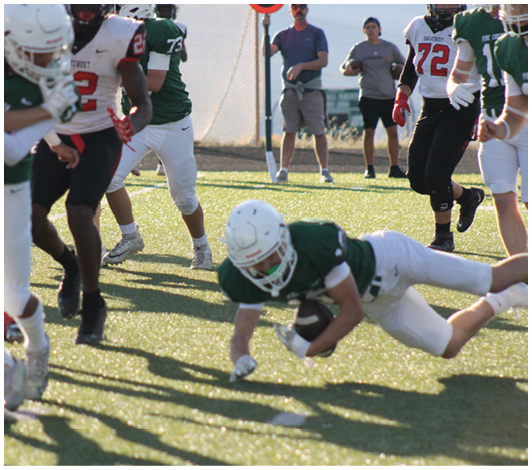 People wearing helmets and sports uniforms are actively playing football on a sunlit field with two people standing in the background. Description generated by AI