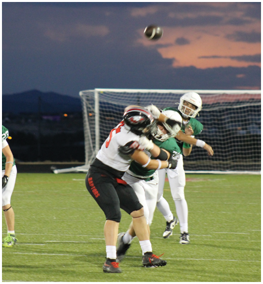 Two people in football uniforms are engaged in a tackle on a field with a goalpost and a football in midair during dusk. Description generated by AI