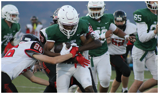 People in football uniforms are engaged in a play on a field with one person holding the ball while others attempt to tackle or block. Description generated by AI