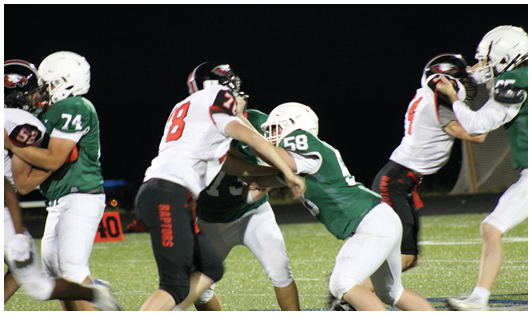 People wearing football uniforms are engaged in a play on a grassy field under stadium lights at night. Description generated by AI