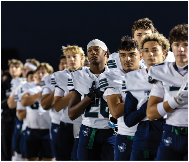 A group of people wearing white sports uniforms with dark stripes stand in a line against a dark background, with one person in the foreground wearing a white head covering. Description generated by AI