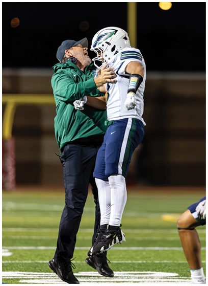 A person in a green jacket and cap is enthusiastically interacting with a football player in white and blue uniform and helmet on a field at night. Description generated by AI