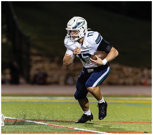 A person wearing a white football helmet and uniform is running while holding a football on a field at night. Description generated by AI