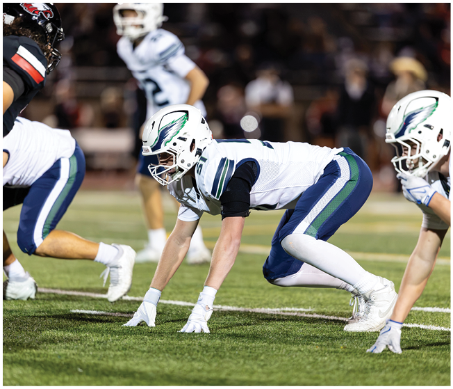 A person in a white football uniform and helmet is crouched in a ready position on a football field with other players in the background. Description generated by AI