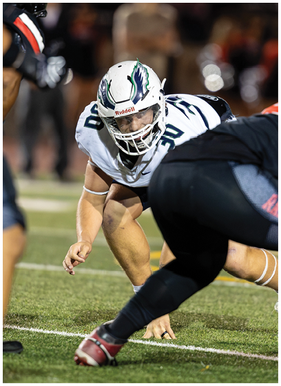 A person in a white football uniform and helmet is crouched on a grassy field facing an opponent in a black uniform during a game. Description generated by AI