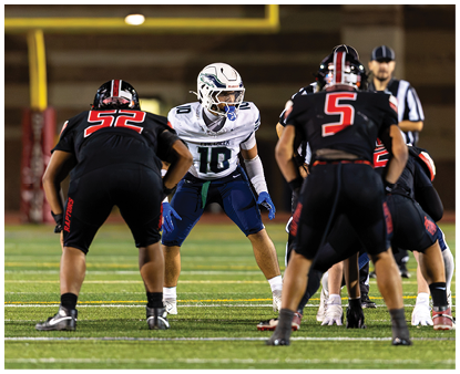 A group of people in football uniforms are positioned on a field, with one person in a white jersey facing two others in black jerseys, preparing for a play. Description generated by AI