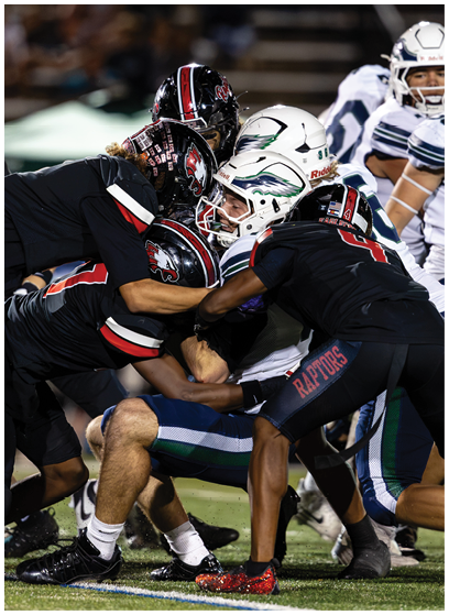 Several football players in dark uniforms tackle a player in a white helmet and uniform during a game on a field. Description generated by AI