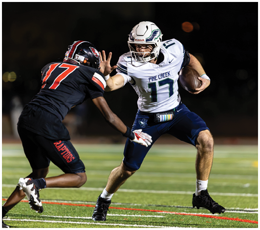 Two people in football uniforms are engaged in a play on a field, with one holding the ball and the other attempting to block or tackle. Description generated by AI