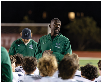 A person in a green football jacket is speaking to a group of people wearing white football uniforms on a field at night. Description generated by AI