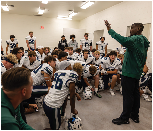 A group of people wearing football uniforms are gathered in a locker room listening attentively to a person in a green jacket who appears to be giving instructions. Description generated by AI
