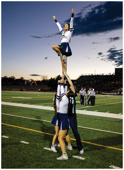 A person in a cheerleading uniform is being lifted into the air by others during an outdoor event at dusk. Description generated by AI