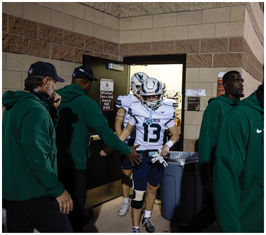 A person in a football uniform with the number 13 is walking out of a doorway while several people in green jackets stand nearby. Description generated by AI
