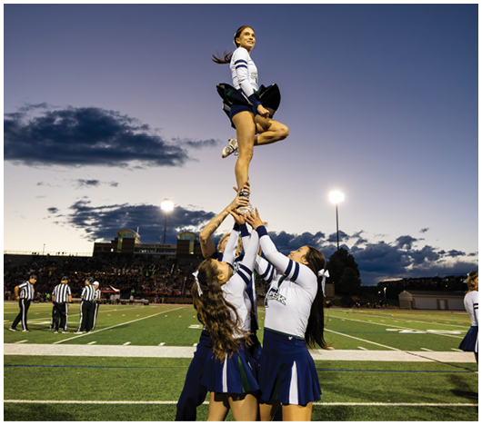 A person in a cheerleading uniform is balanced high in the air supported by the hands of others against a twilight sky with stadium lights in the background. Description generated by AI