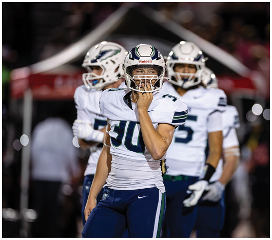 Three people in football uniforms and helmets stand on a field with one person in the foreground holding their faceguard. Description generated by AI