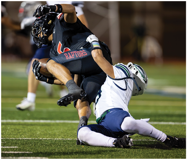 Two people in football gear are engaged in a tackle on a grassy field, with one person in black and red uniform being brought down by another in white and blue uniform. Description generated by AI