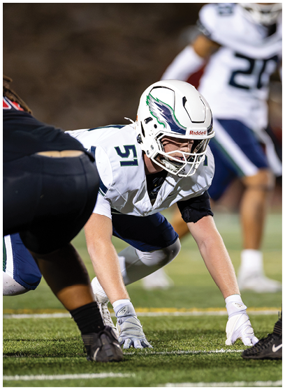 A person in a white football uniform and helmet is crouched on the field in a ready position facing an opponent in a black uniform during a game. Description generated by AI