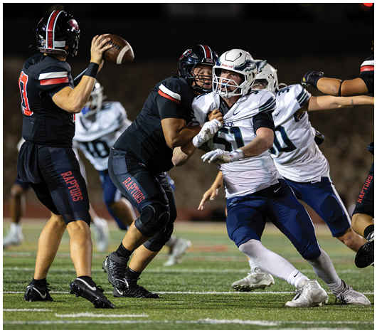 Two football players in black uniforms with “Raptors" on their pants are engaged in a play with one preparing to throw the ball while a player in a white and blue uniform attempts to block or tackle. Description generated by AI