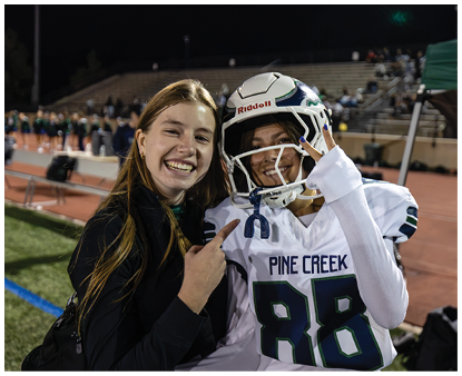 Two people are smiling on a sports field at night, one wearing a football helmet and jersey with the number 88 and the other pointing at them. Description generated by AI