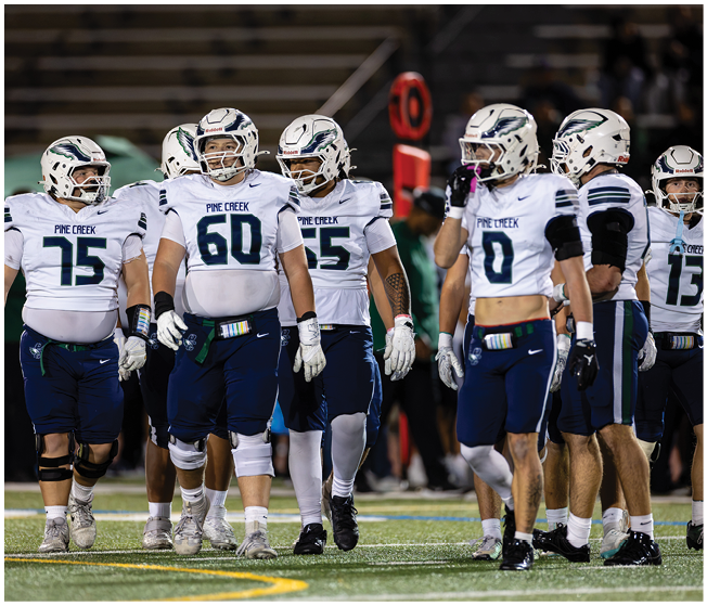 A group of football players in white helmets and navy blue pants walk together on a field during a game. Description generated by AI