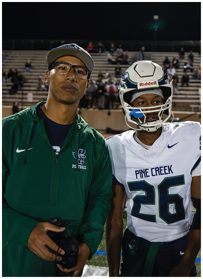 Two people stand side by side on a sports field at night, one wearing a green jacket and glasses and the other in a white football uniform and helmet with the number 26 and “Pine Creek" on the jersey. Description generated by AI
