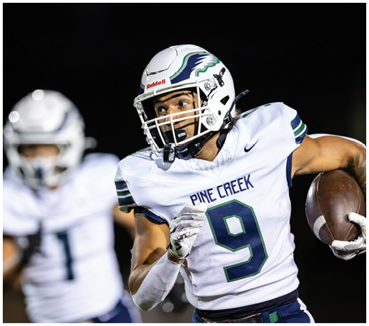 A person wearing a white football uniform with the number nine and a helmet is running with a football while another player in a similar uniform is blurred in the background. Description generated by AI