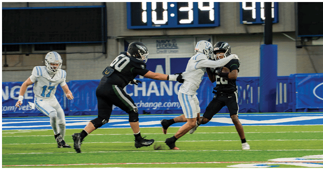 Four people in football uniforms are actively engaged in a play on a green field with a scoreboard and blue banners in the background. Description generated by AI