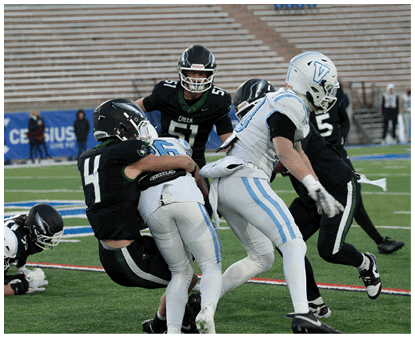 Players in black and white football uniforms are engaged in a tackle on a green field with empty bleachers in the background. Description generated by AI