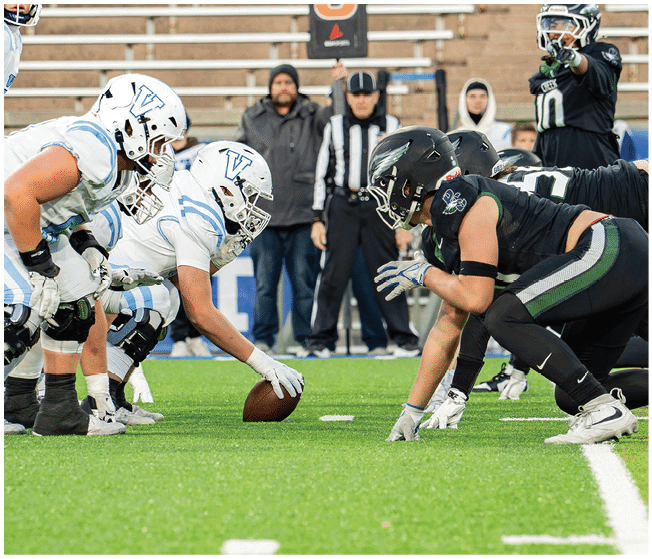 Two opposing American football teams are lined up at the line of scrimmage on a green field with a referee and people in the background. Description generated by AI