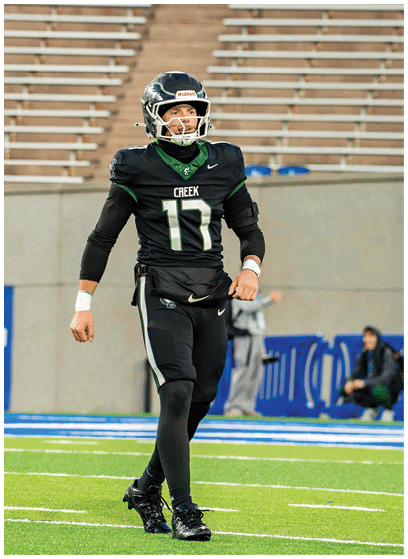 A person wearing a black football uniform with the number 17 and a helmet is walking on a football field with empty bleachers in the background. Description generated by AI