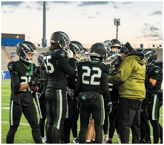 A group of people wearing black football uniforms and helmets are gathered closely on a field while one person in a green jacket and headset appears to be giving instructions. Description generated by AI