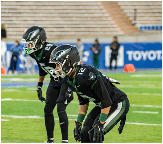 Two people in black football uniforms and helmets are positioned on a green field, appearing ready for a play during a game. Description generated by AI