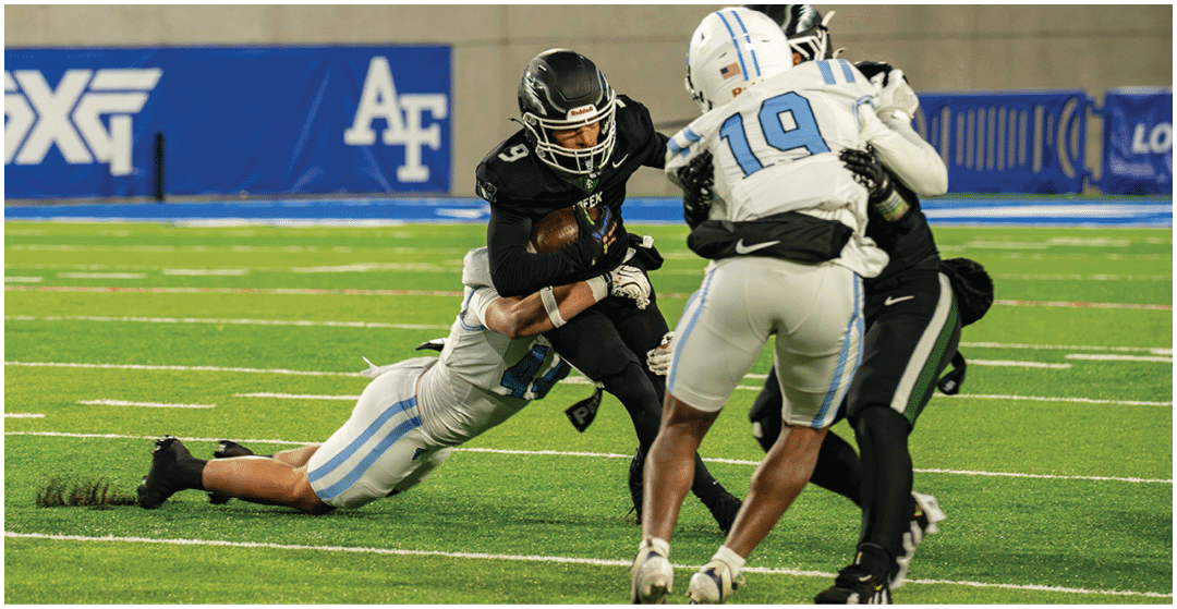 A person in black football gear is running with the ball while being tackled by a person in white and light blue gear on a football field with other players nearby. Description generated by AI