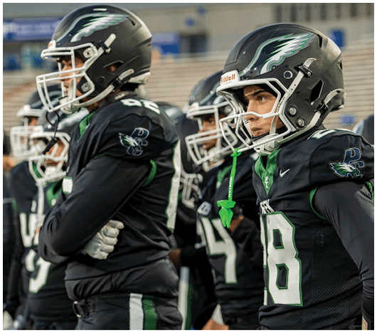 People wearing black football uniforms and helmets stand closely together on a field, appearing focused and ready for a game. Description generated by AI