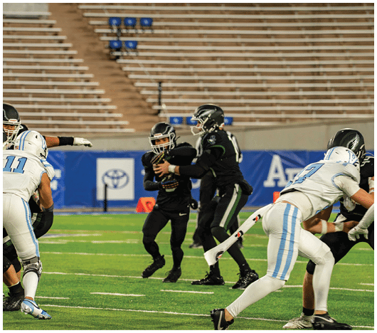 People in football uniforms are actively engaged in a play on a green field with empty bleachers in the background. Description generated by AI
