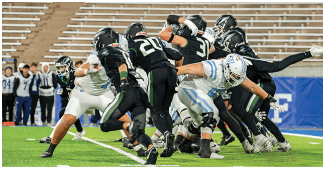 A group of people wearing football uniforms are actively engaged in a play on a green field with empty bleachers in the background. Description generated by AI