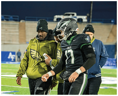 A person wearing a black football uniform and helmet is walking on a field accompanied by two people in green and blue jackets, one holding a small white object. Description generated by AI