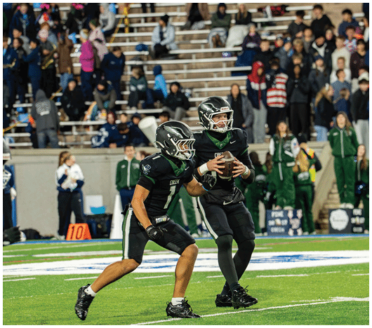 Three football players in black uniforms are on a field with one player holding the ball and preparing to pass while the others move forward, with spectators visible in the background. Description generated by AI