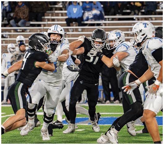 Several people wearing football uniforms are engaged in a physical play on a football field with spectators in the background. Description generated by AI