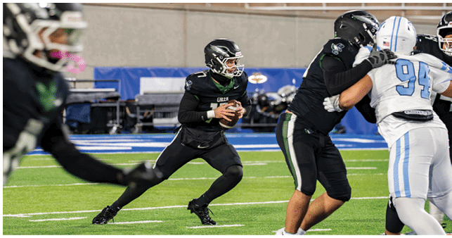A person in black sports gear is holding a football and preparing to throw while other players in black and white uniforms engage on a football field. Description generated by AI
