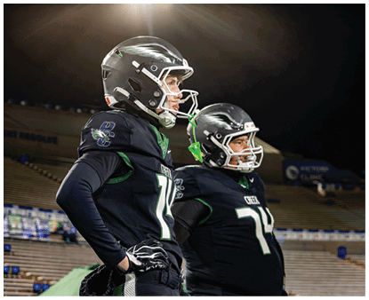 Two people wearing black football uniforms and helmets stand side by side on a field with empty bleachers in the background. Description generated by AI