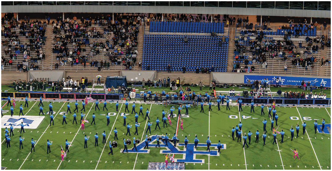 A marching band in blue uniforms is performing on a football field with a large crowd seated in the stadium stands behind them. Description generated by AI