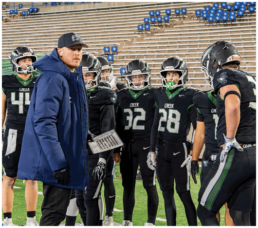 A group of people in football uniforms and helmets stand on a field while one person in a blue coat and cap holds a clipboard and appears to be giving instructions. Description generated by AI