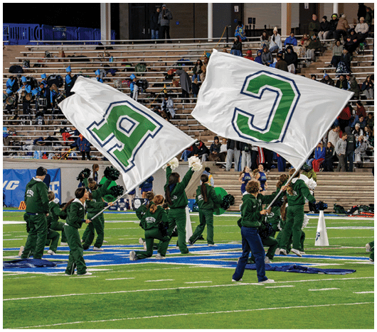 People in green uniforms are on a sports field holding large white flags with green letters while others in the background watch from bleachers. Description generated by AI