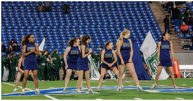 A group of six people in matching blue cheerleading uniforms stand in formation on a football field with blue stadium seats and other people in green uniforms in the background. Description generated by AI