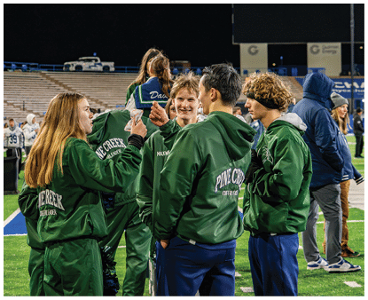 A group of people wearing green jackets with “Pine Creek Cheerleader" on the back are standing together and interacting on a sports field. Description generated by AI