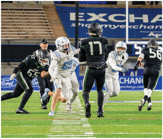 People in football uniforms are actively engaged in a play on a green field with stadium seating and a large advertisement banner in the background. Description generated by AI