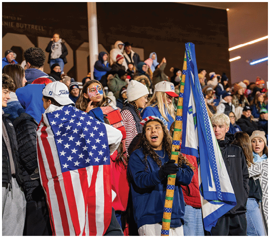 A crowd of people dressed in warm clothing are gathered outdoors, with one person draped in an American flag and another holding a decorated flagpole. Description generated by AI